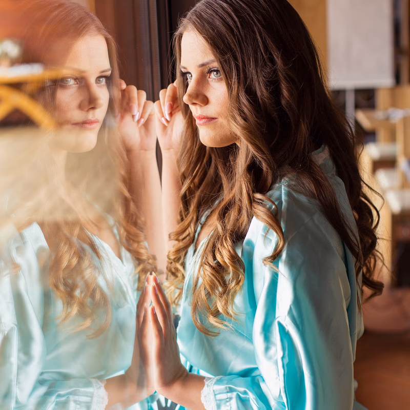 Frau im türkisfarbenen Seidenrobe betrachtet sich nachdenklich im Fenster – Boudoir Shooting Vavrova Photography Göppingen