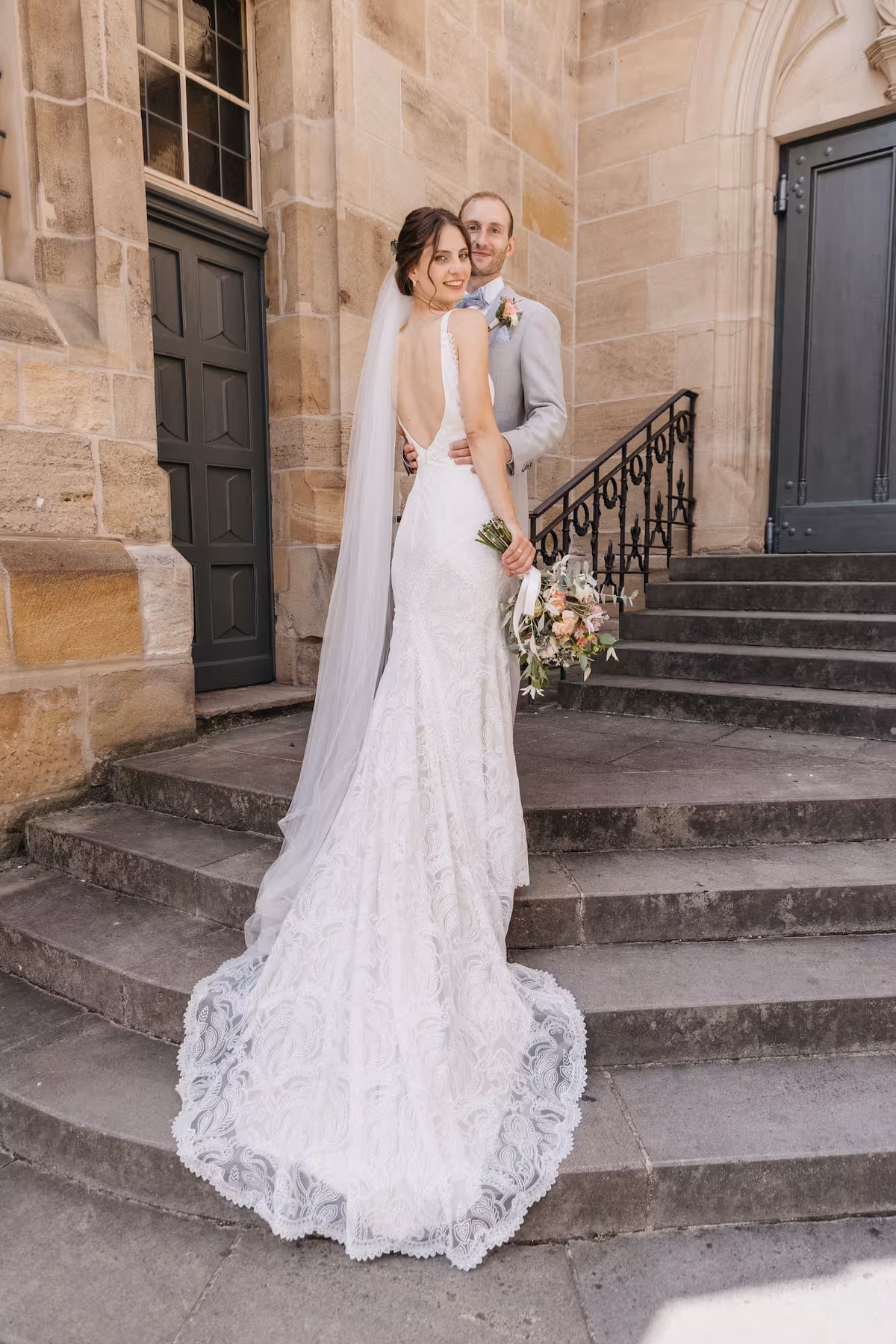 Brautpaar auf historischer Sandstein-Treppe – Braut im eleganten Spitzen-Hochzeitskleid mit Schleier, Bräutigam im grauen Anzug – Hochzeitsreportage Vavrova Photography Göppingen